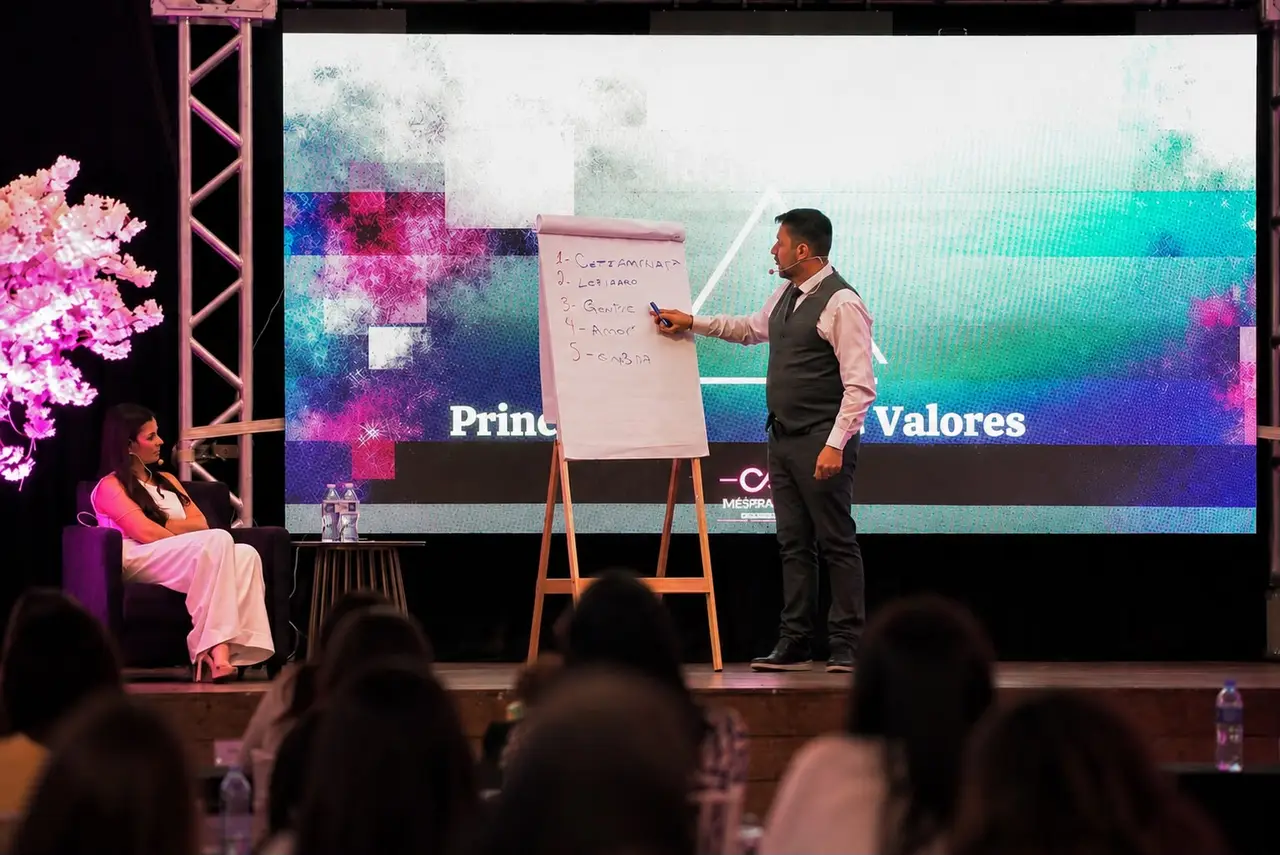 Kim Mattos ensinando em palco de evento, diante de turma e telão